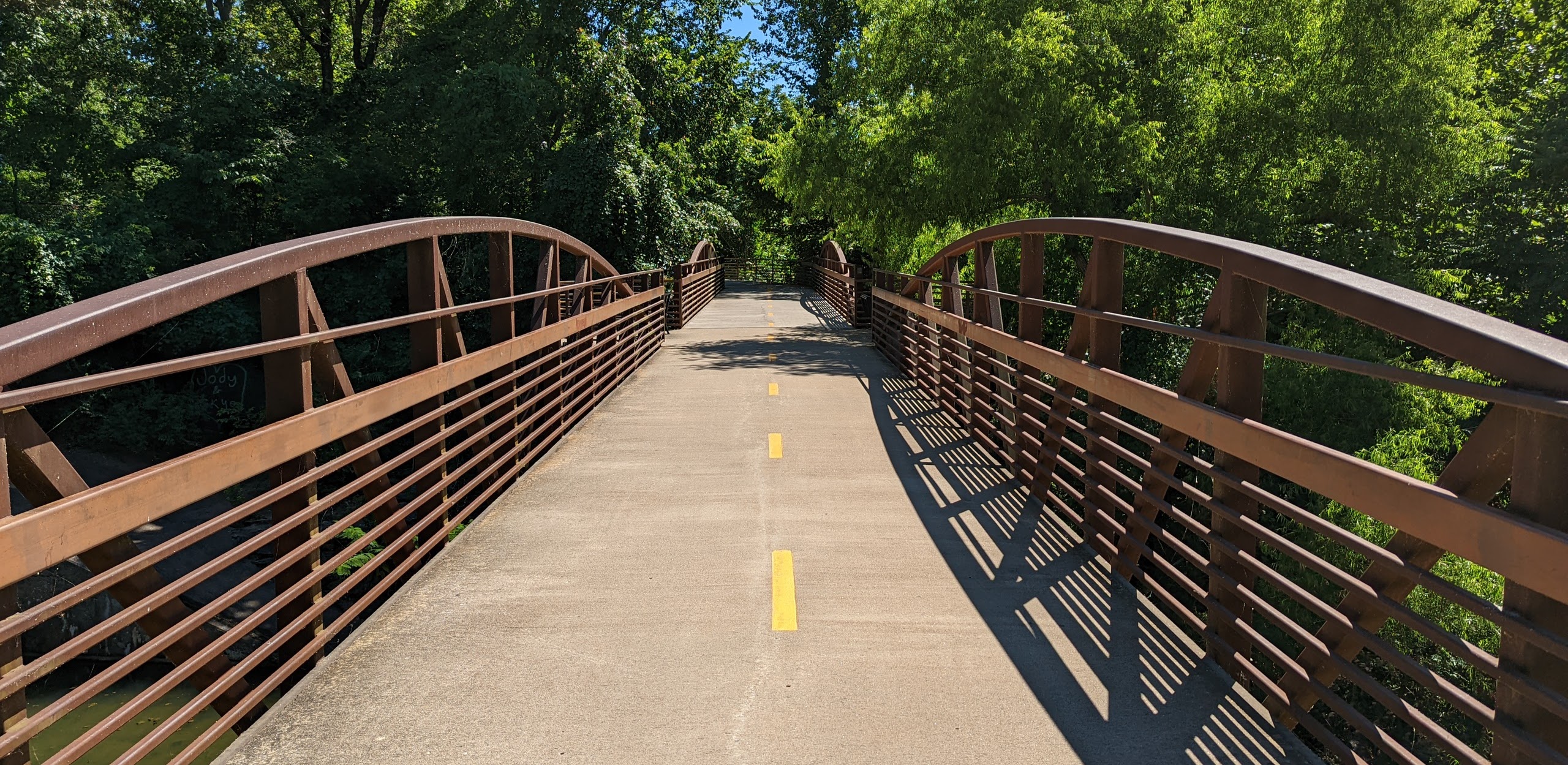 A wide shot of the award winning bridge at fayetteville trail. The bridge has no people on it and the nature in the background is very green. The bridge is paved and has a yellow dotted strip going down the middle of it.