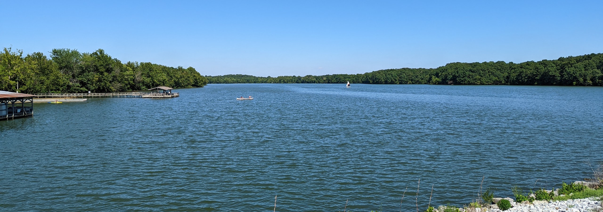 A wide shot of lake fayetteville from the paved trail that surrounds it. There is a canoe and a sail boat in lake fayetteville. It's a bright sunny day and the dock is visible. There a multiple paddle boats visible at the dock.