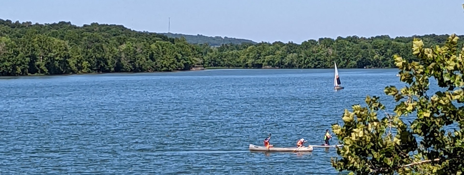 A picture of a sailboat and people in a kayak on Lake Fayetteville. The weather is very sunny and nice and the lake takes up the majority of the picture.