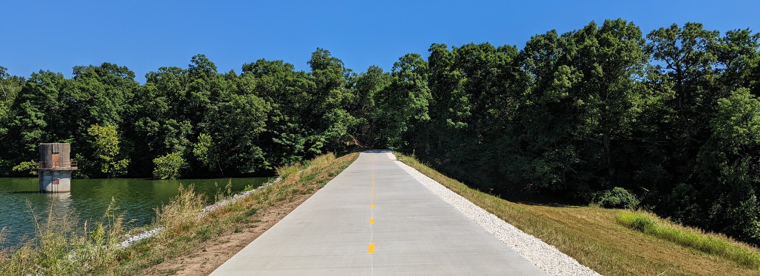 A paved trail that runs along the side of Lake Fayetteville. It's a wide trails with a yellow dotted strip running down the middle of it. It's empty and very sunny outside.