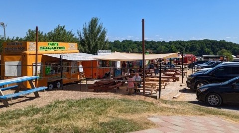 Fabys Mexican food truck which shows a shaded patio in front of the food truck. There is a couple eating and a number of cars parked to the right. Very busy with people in the background.