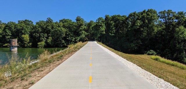 A paved trail that runs along the side of Lake Fayetteville. It's a wide trails with a yellow dotted strip running down the middle of it. It's empty and very sunny outside.