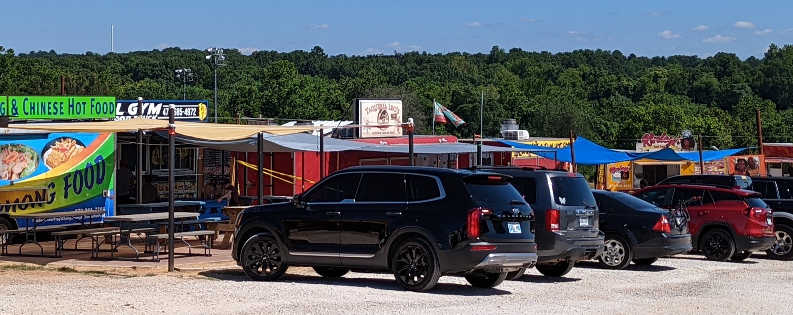 A bunch of cars parked in front of a string of food trucks. It's a bright sunny day and the food trucks are very busy.