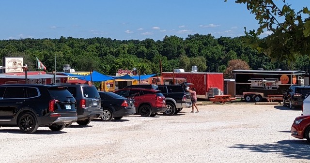 Wider image of the food trucks as a whole. Can see a number of food trucks in the background with car parking in the foreground. It is a very sunny day with lots of commotion in the background near the food trucks.