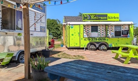An image from a shaded picnic table showing two food trucks up-close in the background. There is an empty picnic table in the foreground.