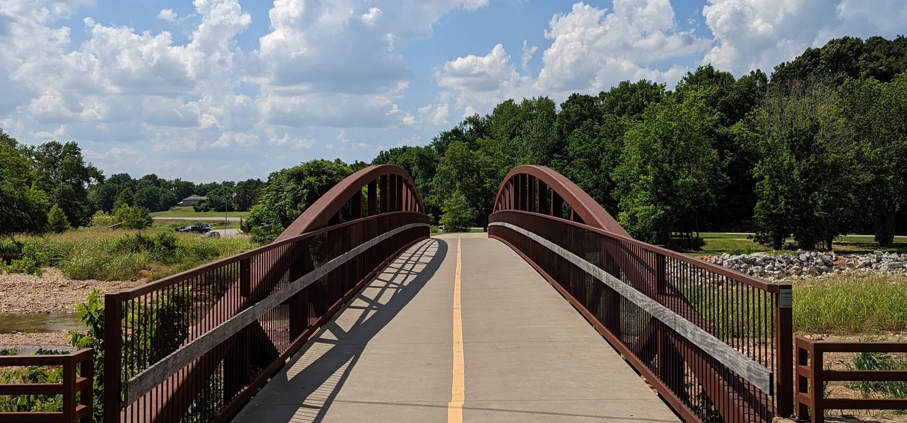 A bridge at JB Hunt Park that is going over a small river/stream. It is very sunny and just a few clouds in the sky.