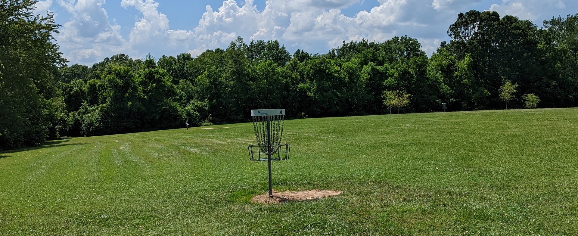 A single disc golf basket in the middle of a field. The field is very green and it's very sunny out.